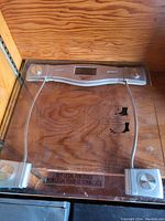 Top-down image of a digital bathroom scale with transparent glass platform and metal circular sensors, digital display in center, placed on wooden surface.