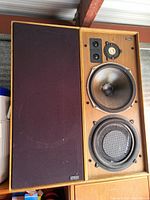 Front view of speaker with fabric grille removed on left side revealing woofer, midrange driver, tweeter and square ports