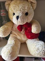 Photo showing a large teddy bear with cream-colored fur, black eyes, and a red bow around its neck.