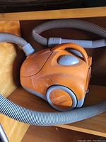 Orange canister vacuum cleaner seen from above with hose coiled around it. Visible scuff marks and dust on body of vacuum.
