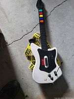 White video game guitar controller on grey floor, showing colored fret buttons, strap, and control knobs.