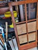 Photo of a medium-tone wooden frame divided into six rectangular sections. The top three sections have glass panes, the bottom three have wooden backing panels. The frame shows wear marks and paint splatters. Background includes garden tools and other household items.