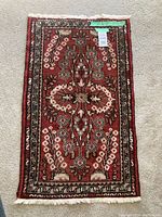 Full rectangular area rug with intricate red, cream, black, and beige floral and geometric pattern lying on carpeted floor.
