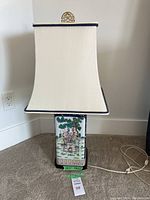 Front view of the lamp showing the cream colored lampshade with blue trim, wooden base, ceramic lamp body decorated with Asian-inspired painted figures and trees.