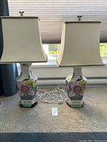 Two matching hexagonal ceramic lamps with multicolor floral designs on white bodies, wooden hexagonal bases, cream lampshades with green edge trim, and white power cords placed in front of a window.