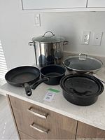 Set of cooking pots and pans: stainless steel stock pot with lid, wok with lid, smaller saucepan, and frying pan on countertop.