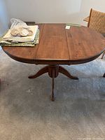 Full view of the wooden pedestal dining table with folded tablecloths and fabric accessories on top, showing the table's round shape and wood finish.