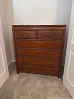 Front view of the wooden tall boy dresser showing six drawers and brass handles, placed on carpeted floor between white doors.
