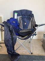 Navy blue and gray folding chair with black carry bag leaning against it, blue G-Tech backpack and gray/blue Tug backpack placed inside the chair.