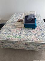 Photo showing queensize mattress with floral pattern and assorted folded bedding on top