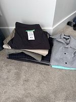Stack of folded men's pants in various colors including dark gray, beige, and plaid patterns alongside folded men's dress shirts in gray and light colors.