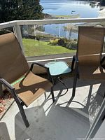 Two brown mesh outdoor chairs and a square glass-top table on balcony with waterfront view.