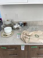 Grouped dinnerware and glassware on counter including Royal Worcester Evesham Gold pieces, Corelle bowls, drinking glasses, and a beige bag.