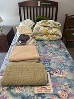 Full view of single bed with mattress, assorted linens folded on top including blankets, sheets, and towels.