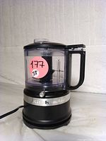 Front view of KitchenAid KFC3516BM matte black mini food processor with clear container and black handle. Pink sticker with number on container.