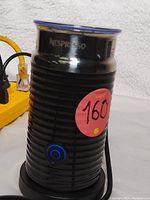 Front view of black Nespresso Aeroccino milk frother with blue lit power button, ribbed design and transparent lid, detailed with red sticker "160" on body.