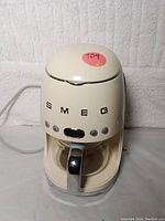 Front view of cream-colored Smeg 50's style programmable drip coffee maker with glass carafe and chrome handle. SMEG brand lettering visible with four control buttons and digital display.