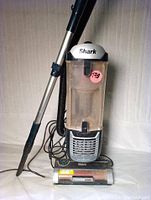 Full view of Shark vacuum upright with metal extension wand, hose, and power cord coiled at base. Clear dust bin is attached showing dust inside. Unit has Shark branding on top.