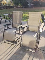 Two beige zero gravity patio chairs with black laced sides and green plastic armrests sitting outdoors on concrete.
