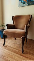 Front right angle view of antique tan wingback chair showing fabric upholstery, wooden arm and leg details, set on hardwood floor near wall.