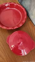 Top down view of red ceramic pie plate with ruffled edges and red petite maison mini lidded baker with lid side by side on wooden surface