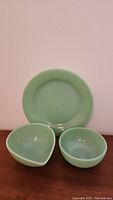Jadeite green Fire King dinner plate, round bowl, and pouring bowl shown together on wood surface against pink wall.