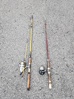 Two fishing rods placed side by side on pavement, one yellow with attached reel and the other red with cork handle and attached reel.