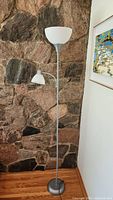 Full view of the two-headed floor lamp standing against a stone wall on a wooden floor.