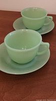 Two Fire King jadeite cups with matching saucers displayed on a wooden surface with visible ribbed pattern on cups and smooth saucers.