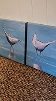 Pair of square canvas prints showing white birds standing on water with reflections, on blue backgrounds, oriented side by side.