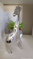 Front angle view of polished aluminum giraffe sculpture on shelf with small plant in background.