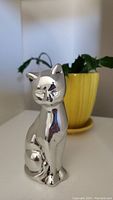 Front view of a polished aluminum cat sculpture showing detailed face and posture, placed against a background with a yellow pot.