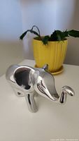 Polished aluminum charging elephant sculpture with raised trunk shown alongside a yellow pot plant for scale.