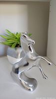 Front angled view of polished aluminum horse sculpture with a potted plant in background.