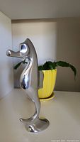 Side view of polished aluminum seahorse sculpture with yellow potted plant in background showing the full shape and clean reflective surface.