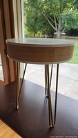 Round accent table showing white round top with jute trim on sides and gold metal hairpin legs in front of window