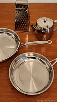 Photo showing two stainless steel frying pans, a rounded teapot with a lid, and a metal box grater arranged on a wooden surface.