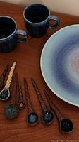 Photo showing two dark blue vintage ceramic mugs with handles and a large round ceramic plate with pink and blue glazing. Seven handcrafted ceramic spoons with twisted handles in various colors are arranged beside them.