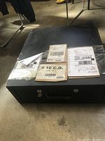 Front and top view of the black metal cash box showing lock, handle and multiple shipping labels on top.