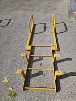 Top-down view of yellow metal loading dock ladder on pavement, showing three rungs and mounting brackets.