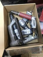 Top-view photo of a box containing a variety of used air tools: nail guns, grinder, pneumatic stapler or nailer, all metal with signs of wear and usage.