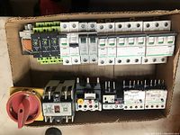 Photo showing a cardboard box with various motor control equipment including circuit breakers, starters, a red/yellow rotary switch, and a thermal overload relay, all arranged in two rows.