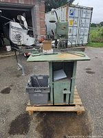 Punch press machine and parts bin positioned on wooden pallet outside near shipping container. Machine shows surface wear.