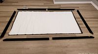 Assembled view showing the unfolded projector screen fabric laid flat inside the unassembled black frame pieces on a wood floor.
