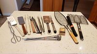 Wide view of most BBQ tools arranged on a kitchen countertop showing different spatulas, tongs, skewers, and grilling racks.