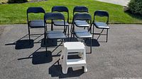 Six black metal folding chairs with vinyl-covered foam seats arranged outside with one white plastic two-step stool in front.