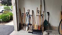 Wide shot showing all garden and cleaning tools stored upright against a wall in garage, including shovels, rakes, brooms, spades, and a black snow shovel.