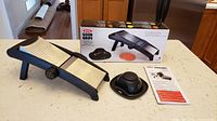 Mandoline slicer set on countertop with food holder, instruction manual, and box in background.