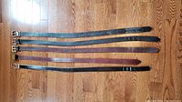 Five leather belts laid out flat on wood floor, showing length and colors
