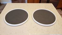 Top view of two round white-framed Lazy Susans with black textured non-skid surfaces on a beige kitchen countertop.